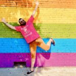 Nick P., the author of swellmode.lol, standing on one leg in front of a brick wall with a large pride flag painted on it.
