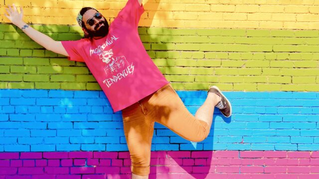 Nick P., the author of swellmode.lol, standing on one leg in front of a brick wall with a large pride flag painted on it.